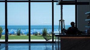 Ein Zimmer mit großen Fenstern mit Blick auf einen Strand, ein Sessel und ein Schreibtisch mit einem Laptop.
