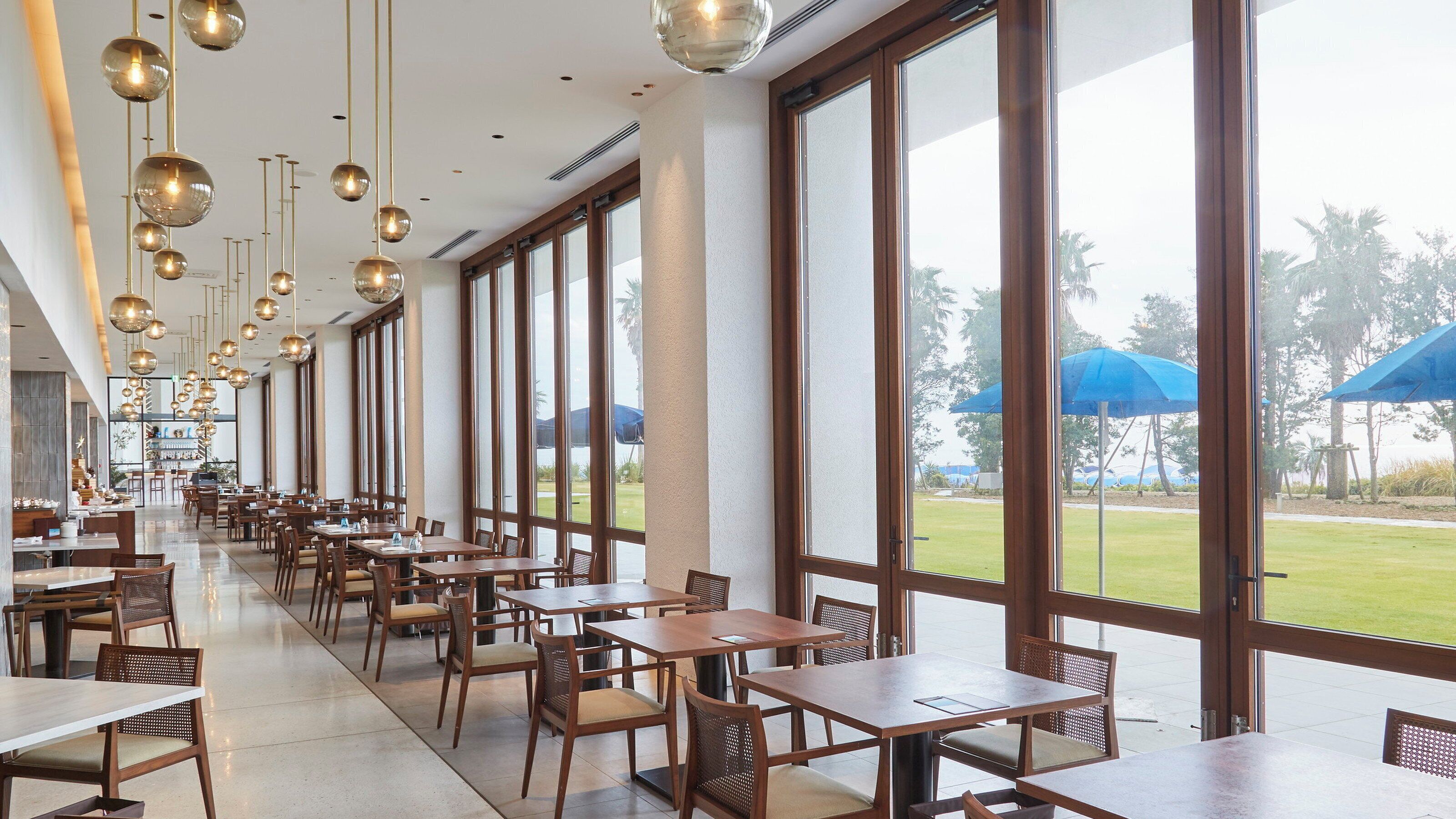A spacious indoor dining area with large windows, wooden tables, and hanging spherical light fixtures.