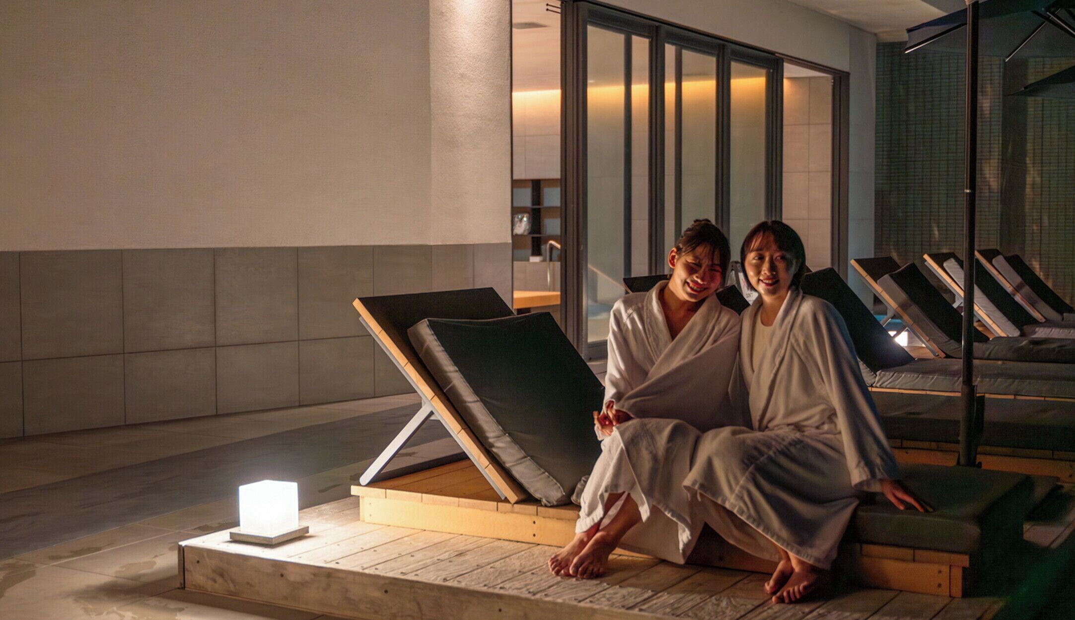 Two people sitting on a wooden platform in a spa area with massage chairs in the background.