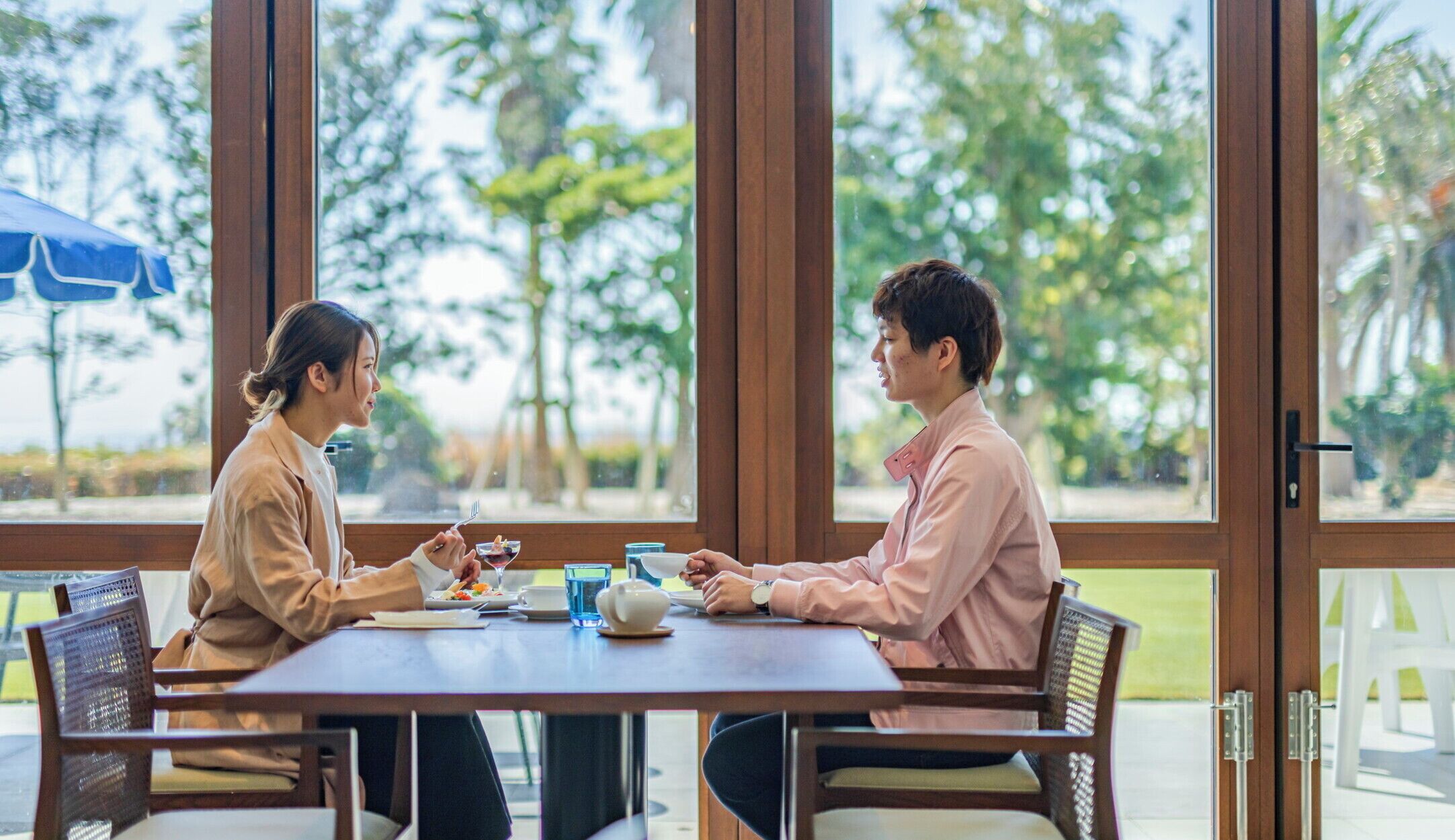 Dos personas sentadas en una mesa de madera con vistas a árboles a través de amplias ventanas.