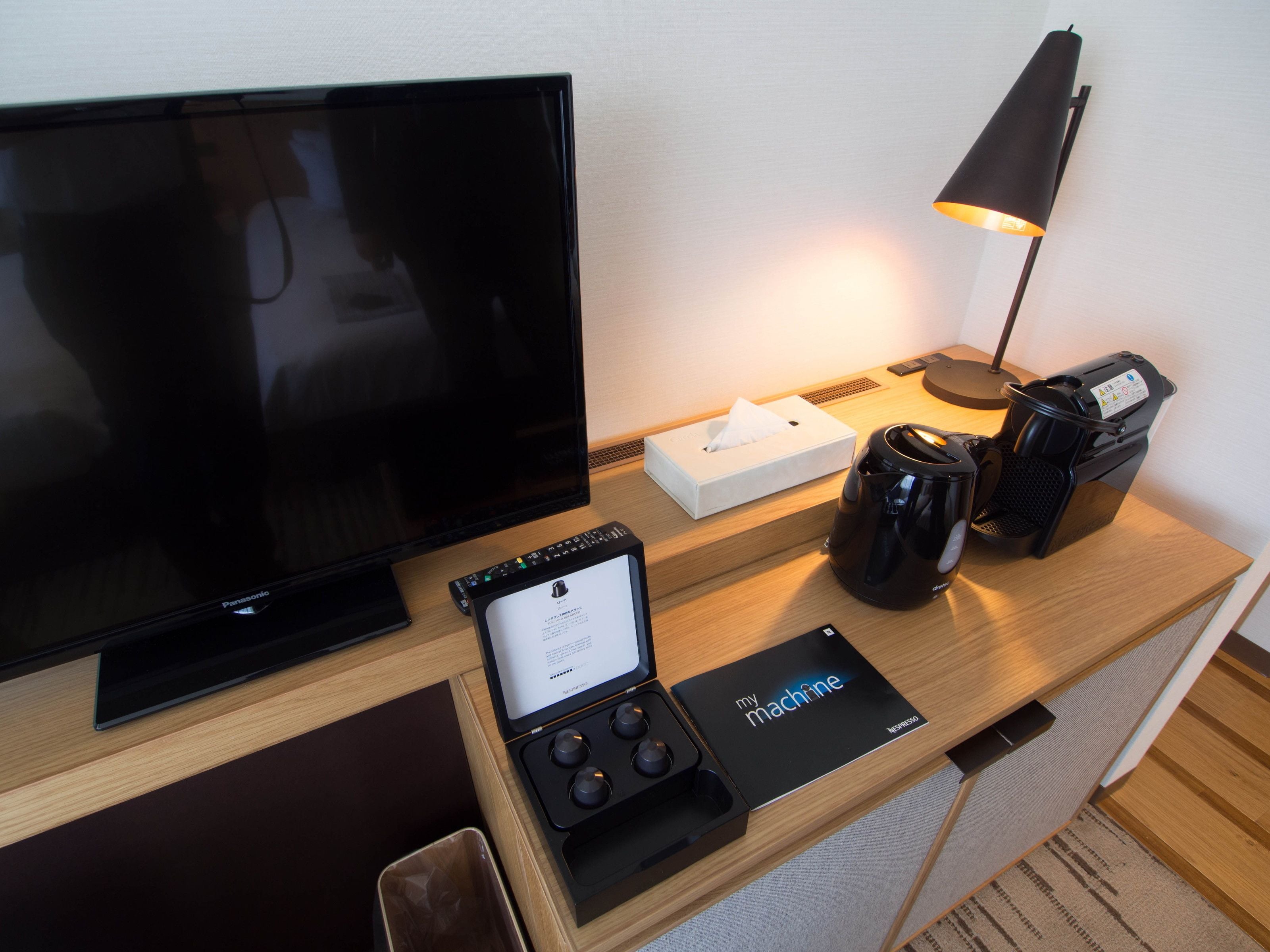 A hotel room with a flat-screen TV, a coffee maker, a lamp, a tissue box, and a remote control.