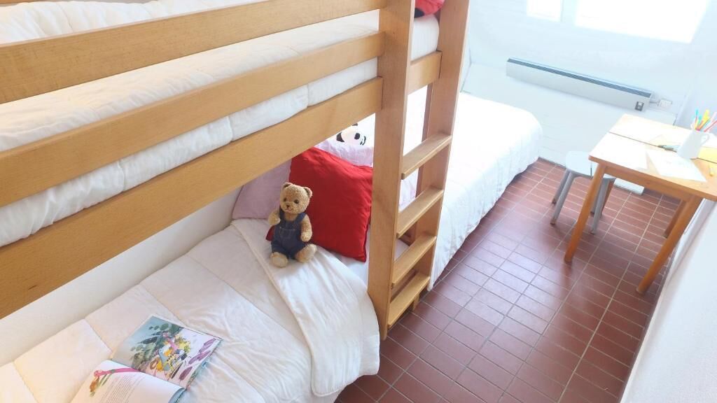 A bunk bed with a teddy bear on the lower bunk, a red pillow, and a small table with a book and a cup.