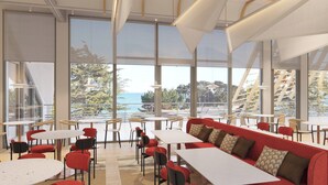 Un comedor moderno con mesas y sillas, ventanas amplias con vista al océano y árboles, y un banco con cojín rojo.