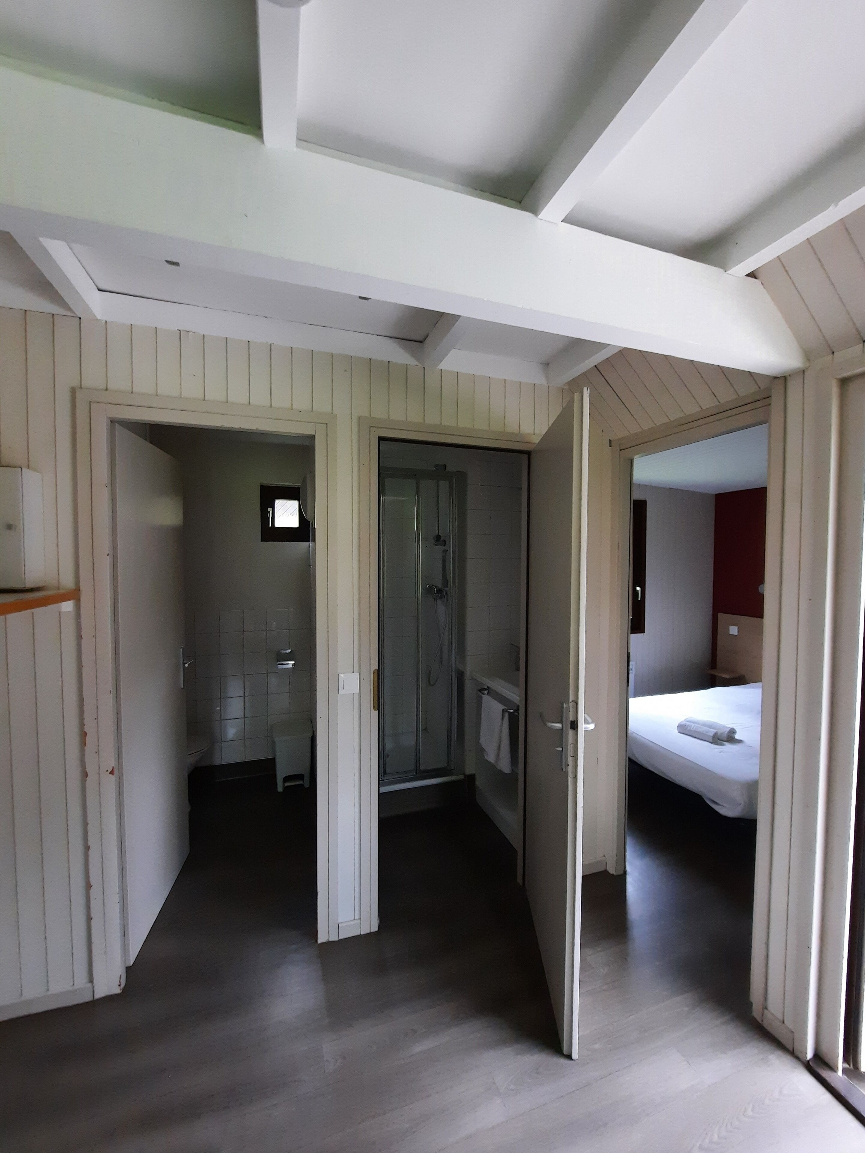 A hotel room with a wooden floor, a shower, a bed, and a bathroom with a toilet and sink.