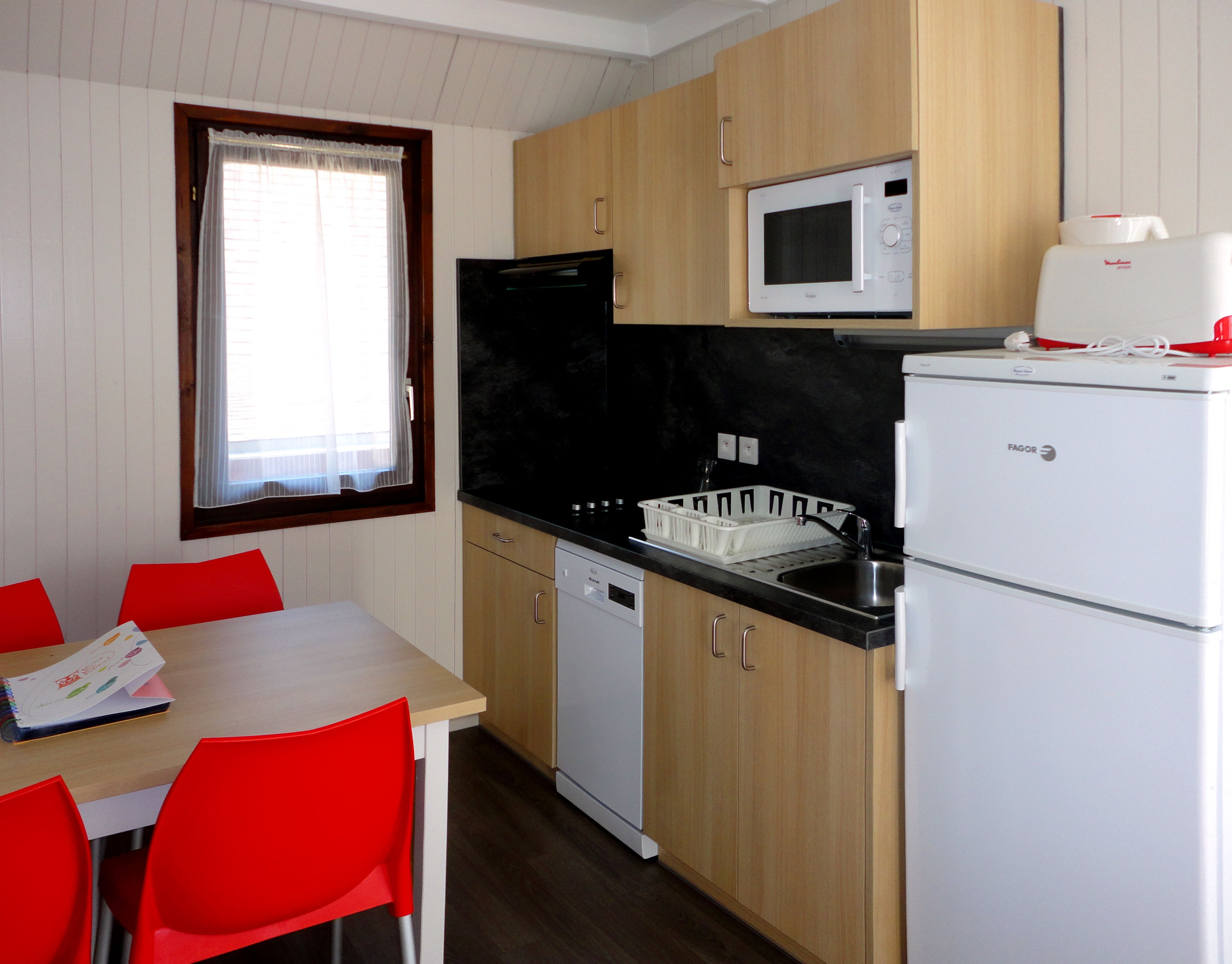 A compact kitchen with wooden cabinets, a black countertop, a white microwave, a white refrigerator, and a window with a white curtain.