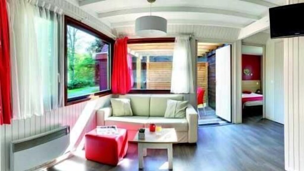 A modern room with a sofa, a small table, and a red ottoman. There is a window with curtains, a door leading to another room, and a television mounted on the wall. - VVF Les Vosges Bitche (Bitche)