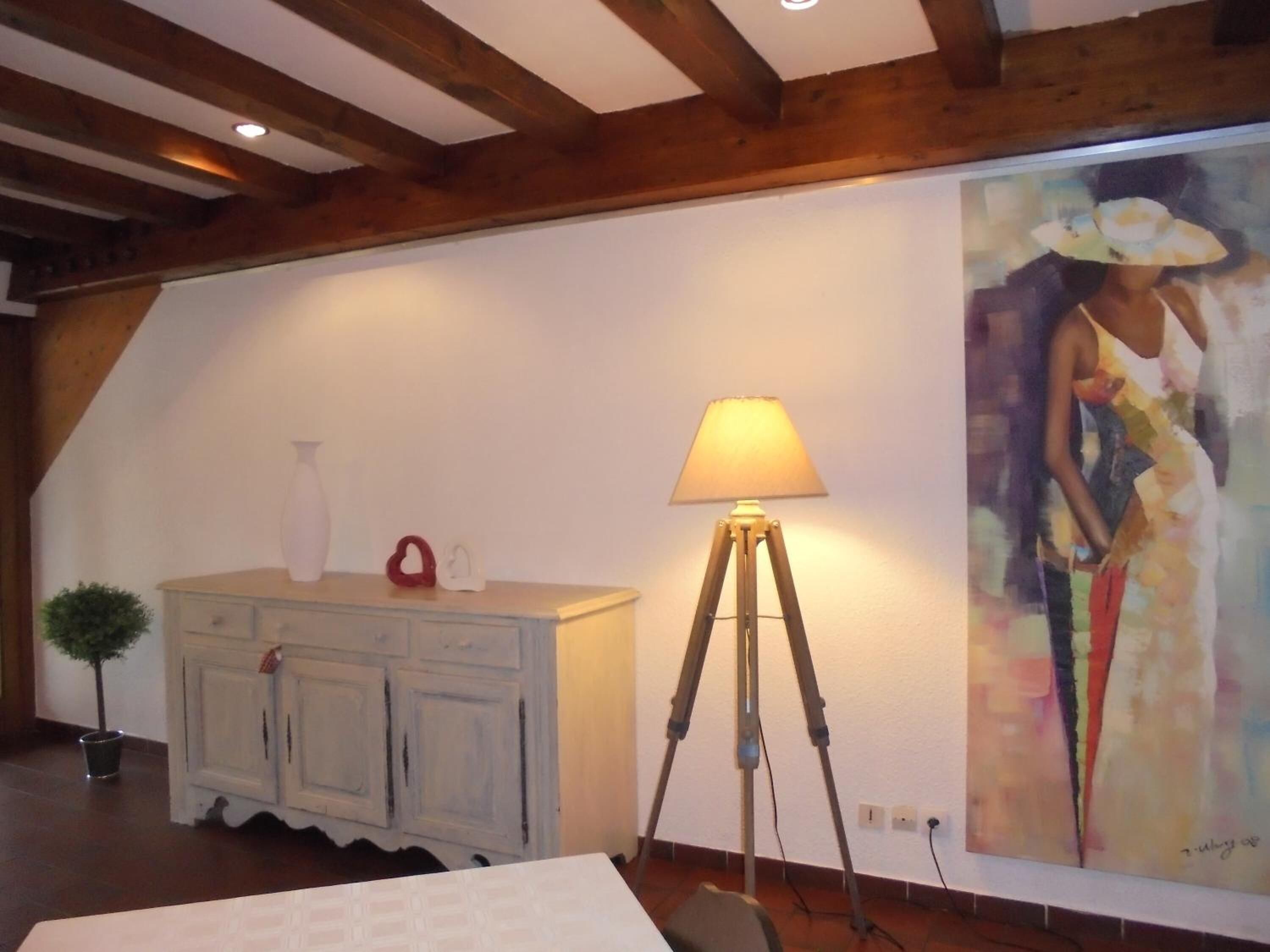 A room with a wooden ceiling, a white cabinet with decorative items, a floor lamp, and a large painting on the wall.