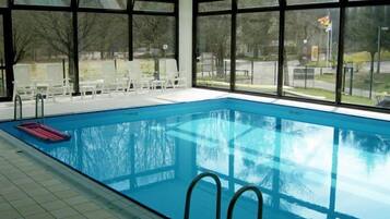 An indoor swimming pool with clear glass walls, a tiled floor, and a few lounge chairs outside.