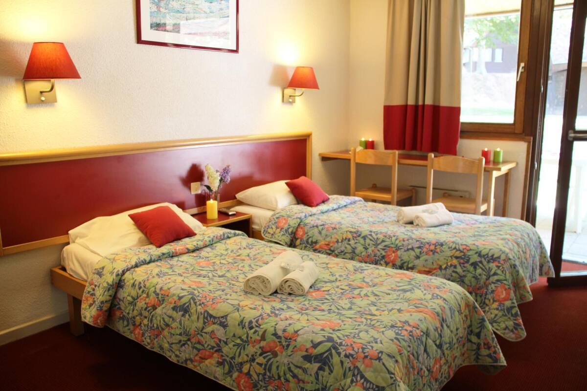 Camera d'albergo con due letti, ognuno con lenzuola a motivi floreali e cuscini rossi. Sono presenti lampade a parete, un tavolino con una candela e una finestra con tende.