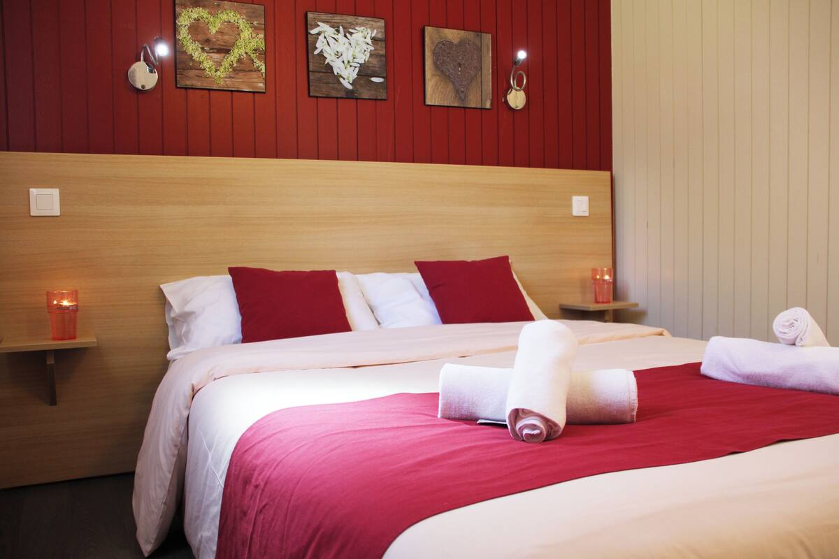 Camera d'albergo con testiera in legno, biancheria rossa e bianca, due comodini con candele e un'opera d'arte appesa al muro.