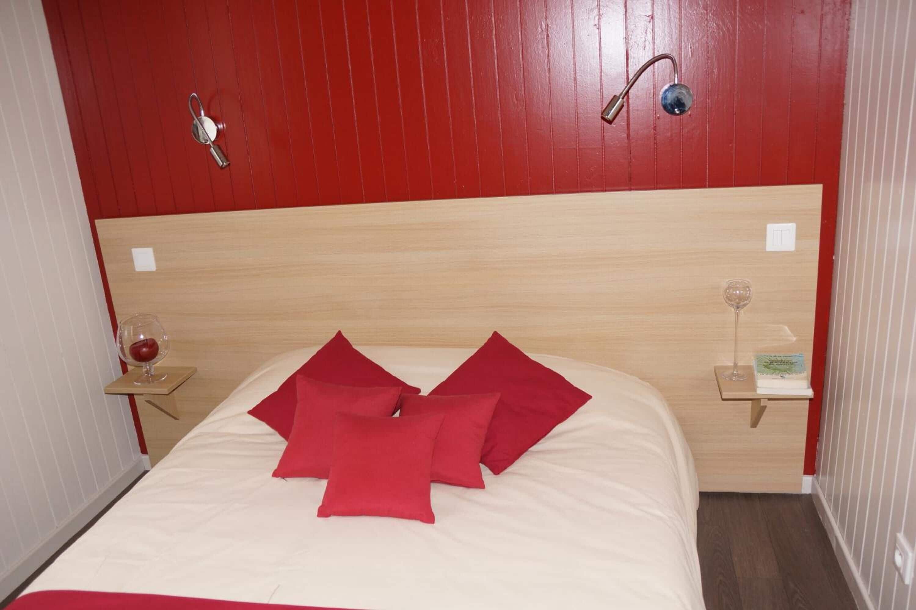 Une chambre d’hôtel avec une tête de lit en bois, un lit avec des oreillers rouges, une table de chevet avec un verre à vin et une petite table de nuit avec une lampe.