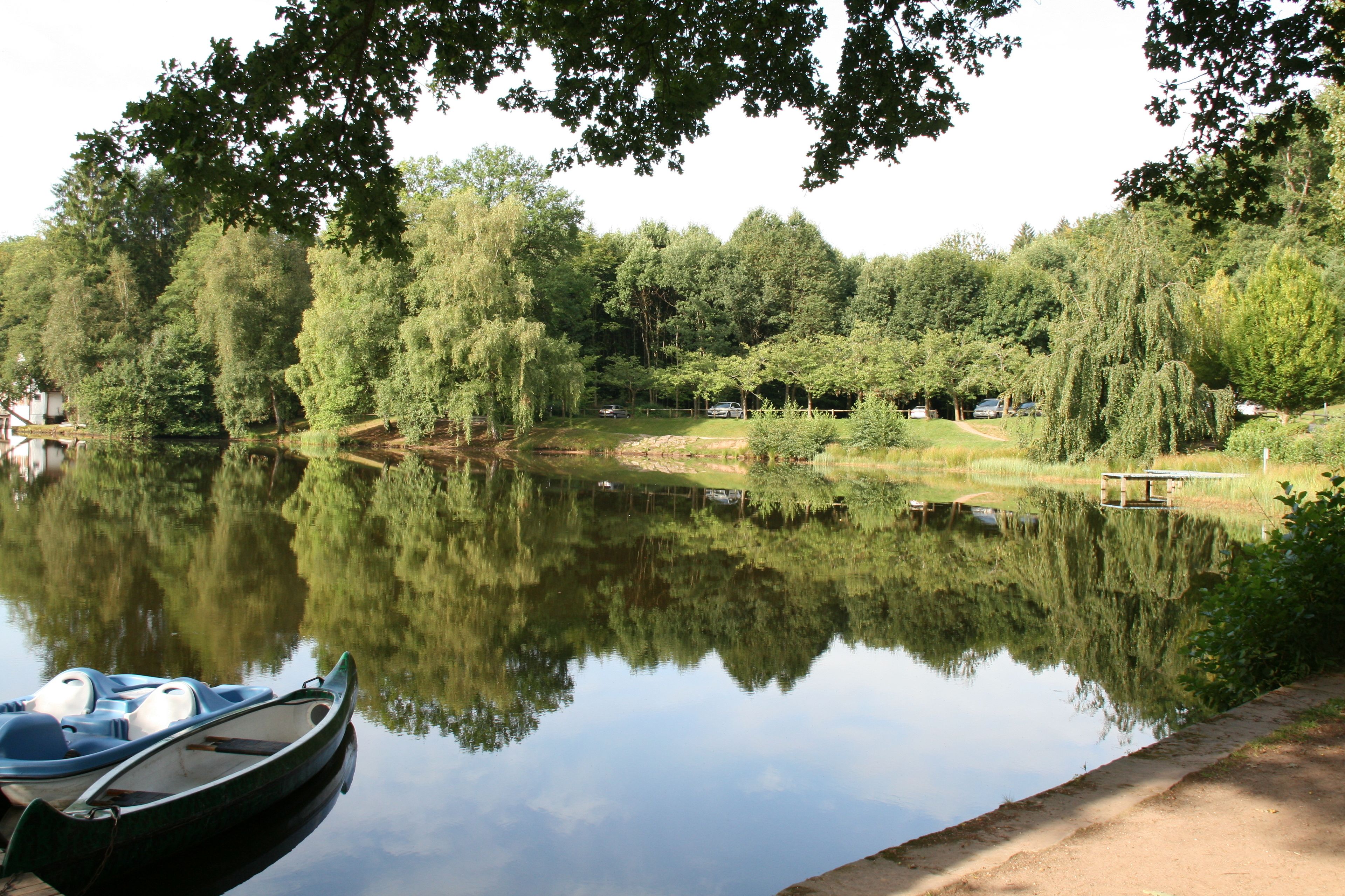 Un lac paisible, avec un bateau amarré sur la rive gauche, entouré d’une végétation luxuriante et d’arbres.