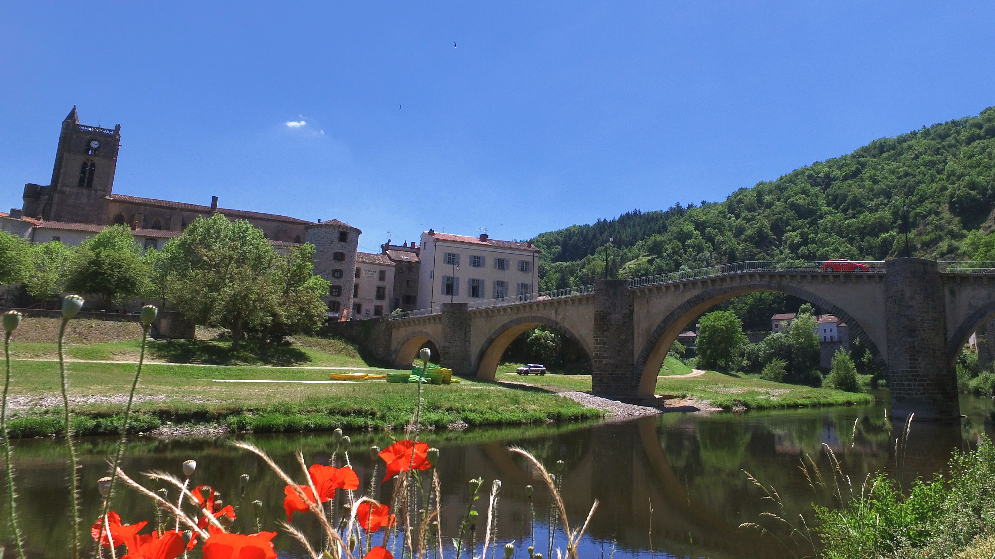 Un pont de pierre enjambant une rivière, avec une ville en arrière-plan, dont on aperçoit le clocher d’une église.