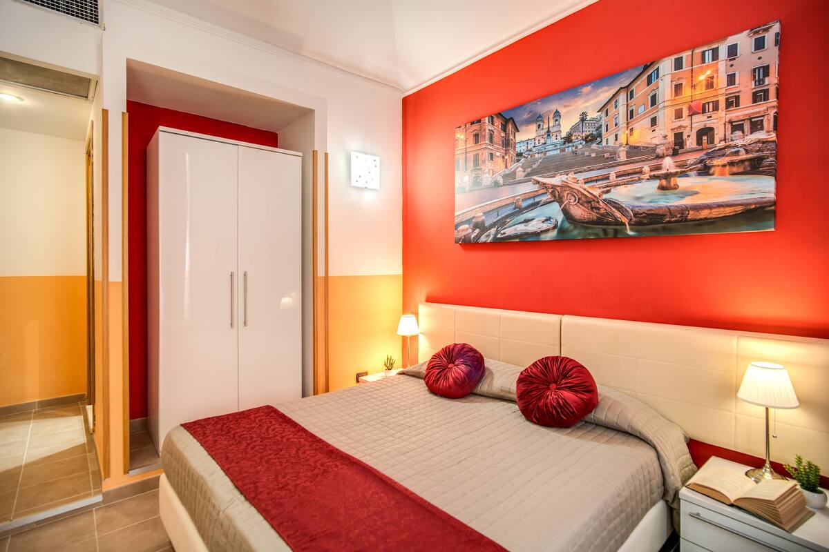 Habitación de hotel con cama, pared roja, un cuadro y mesita de noche con lámpara.