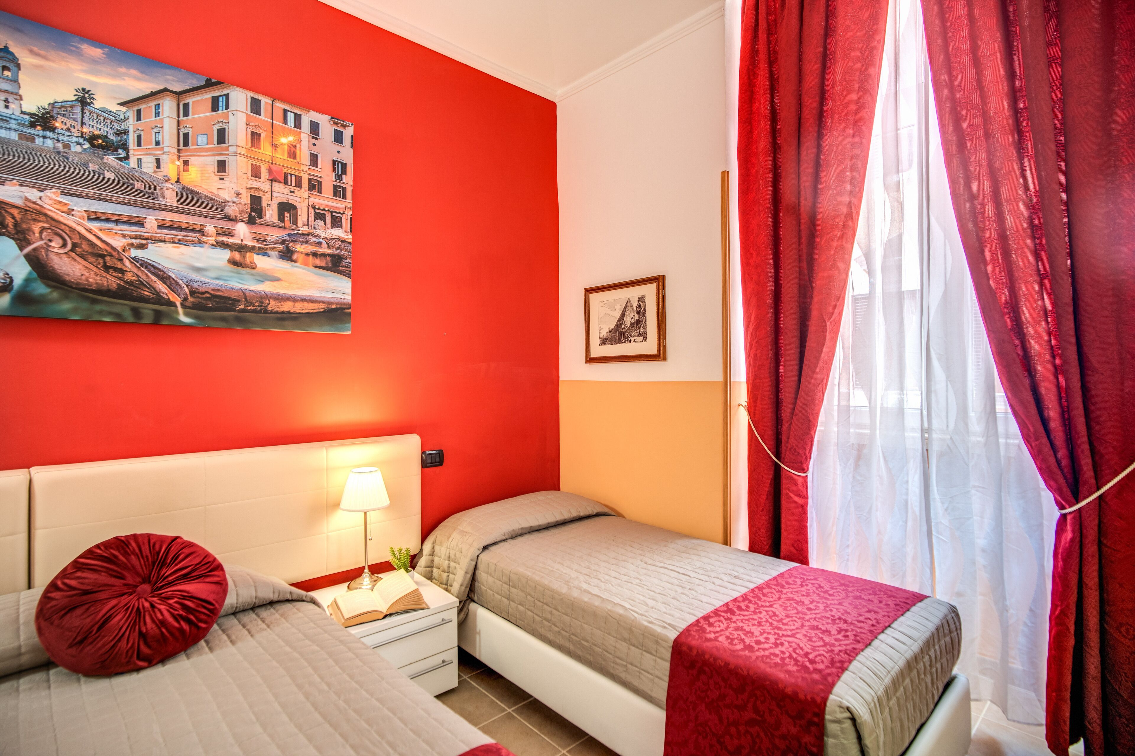 Une chambre d’hôtel avec un mur d’accent rouge, un lit avec un oreiller rouge, une table de chevet avec une lampe et un tableau représentant un paysage urbain.