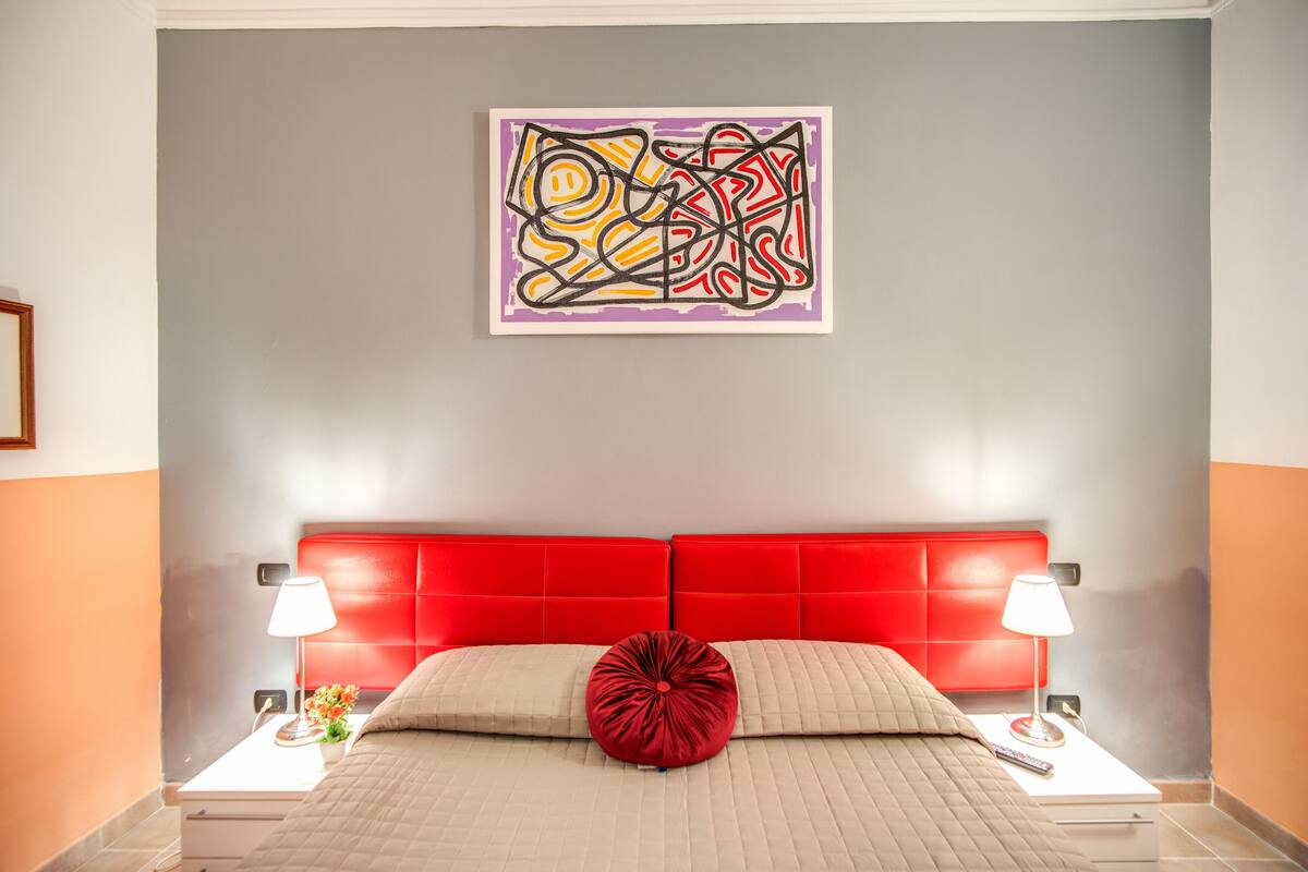 Una habitación de hotel moderna con un cabecero tapizado rojo, una colcha color beige y una almohada decorativa roja. Se observa una obra de arte abstracta colgada en la pared sobre la cama.