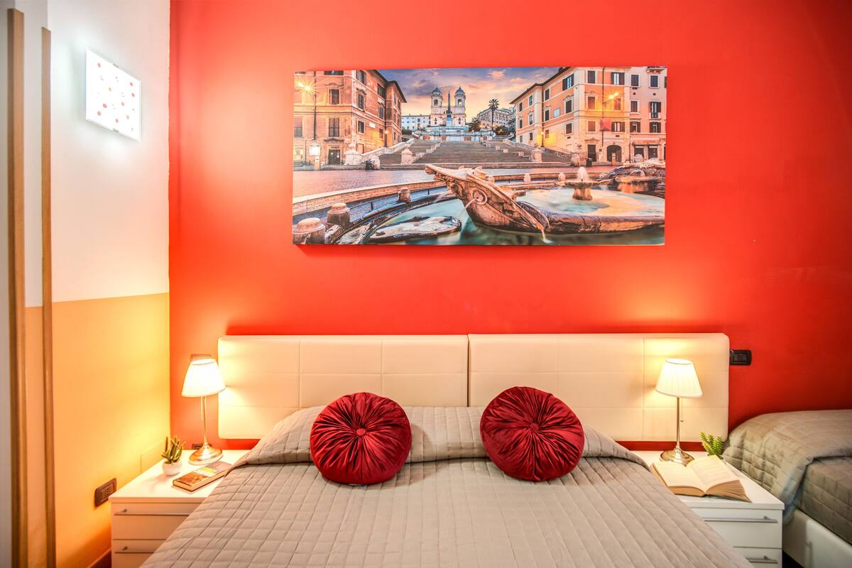 Una habitación de hotel con una pared roja, una cama con dos almohadas rojas, una mesita de noche con una lámpara y un cuadro de un paisaje urbano.