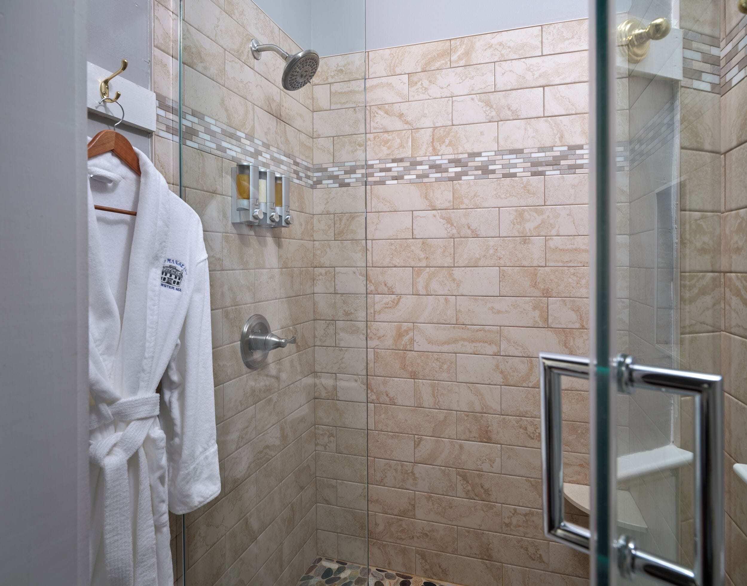 A glass-enclosed shower with beige tiles, a white robe hanging on a hook, and a glass door.