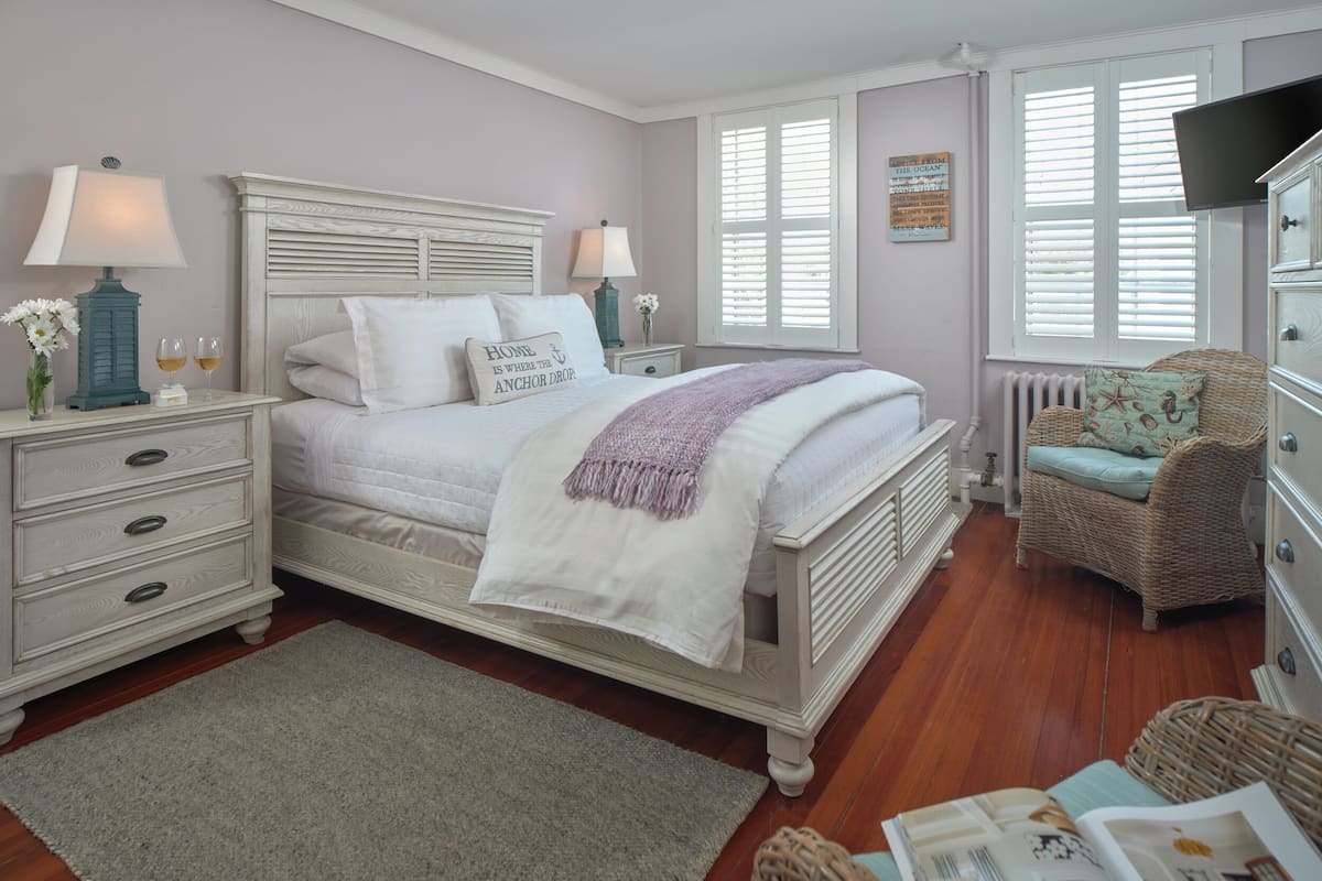 Une chambre à coucher comprenant un lit, des tables de chevet, une commode, un fauteuil en osier et une fenêtre avec des stores.