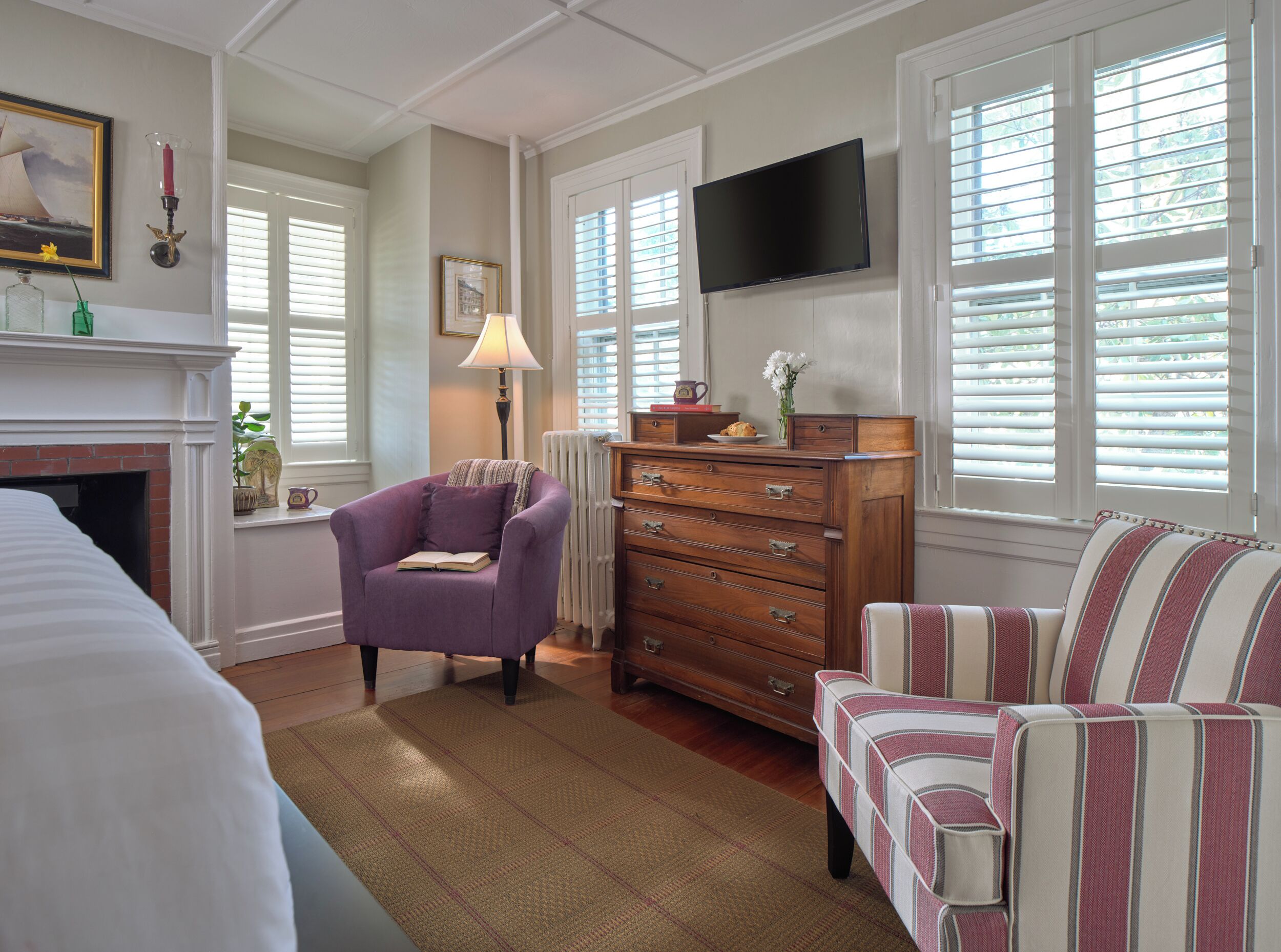 Une pièce avec une cheminée, un téléviseur fixé au mur, un fauteuil violet, un canapé à rayures et une commode en bois.