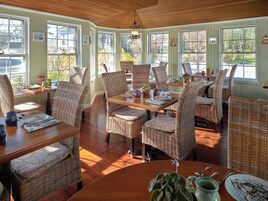 Un comedor con mobiliario de mimbre, mesas de madera y grandes ventanales con vistas a árboles.