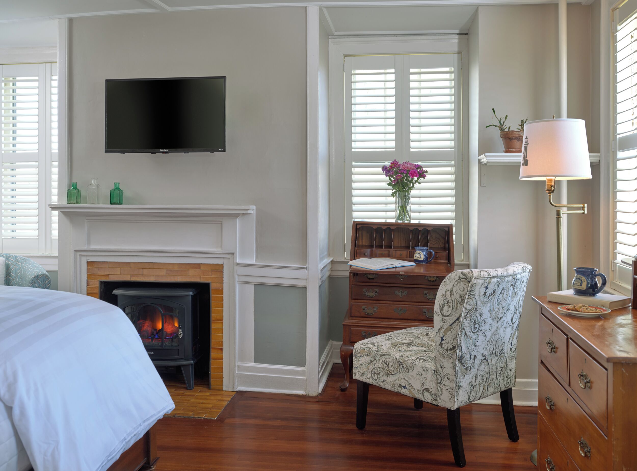 A room with a fireplace, a television mounted on the wall, a desk with a lamp, and a chair.