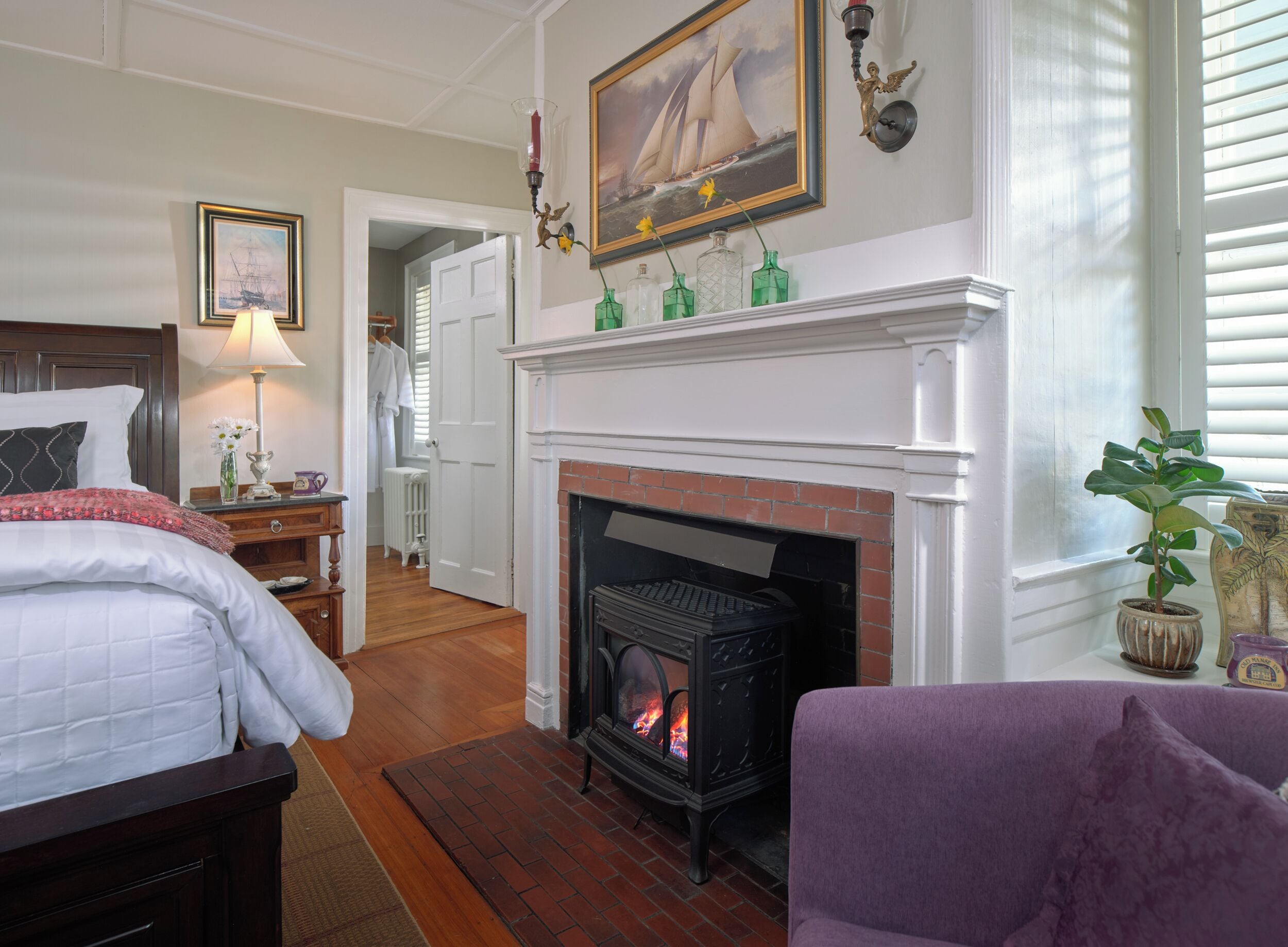 Una habitación de hotel con chimenea, una cama con ropa blanca, una mesita de noche con lámpara y un pasillo que conduce a otra habitación.