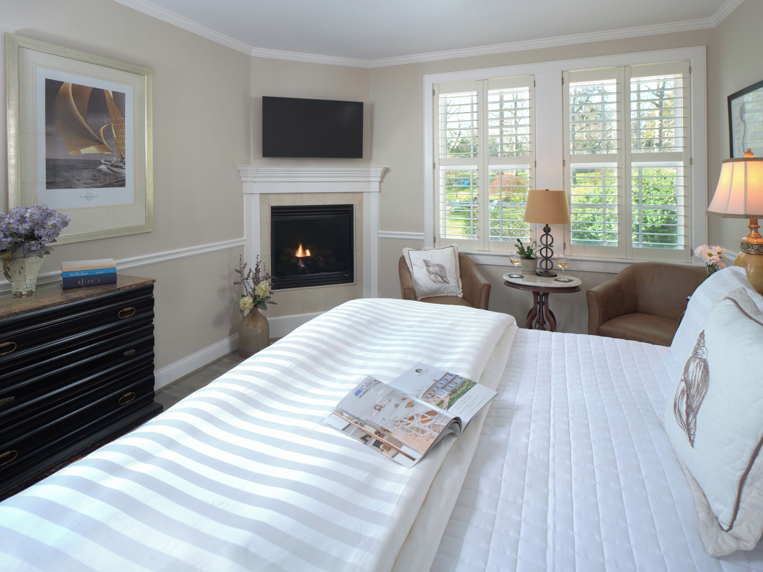 A hotel room with a fireplace, a bed with a magazine, a desk with a lamp, and a window with a view of greenery.