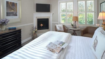 A hotel room with a fireplace, a bed with a magazine, a desk with a lamp, and a window with a view of greenery.