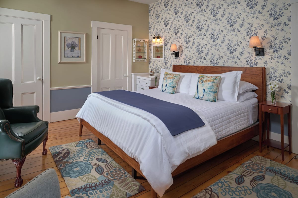 Une chambre à coucher avec un lit en bois, du linge de lit blanc, un papier peint à motifs floraux, un fauteuil vert et un tapis à motifs.