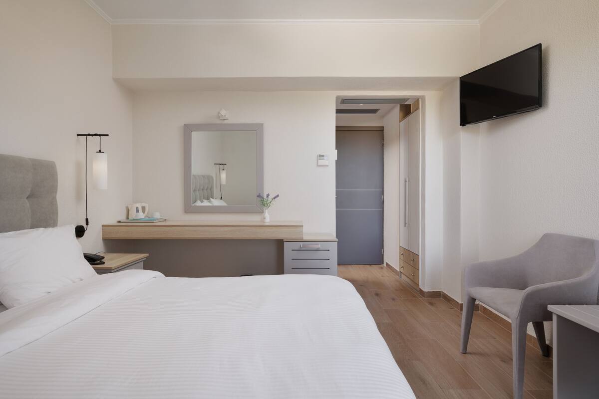 A modern hotel room with a large bed, a bedside table, a flat-screen TV mounted on the wall, a small desk with a chair, and a bathroom visible through an open door.