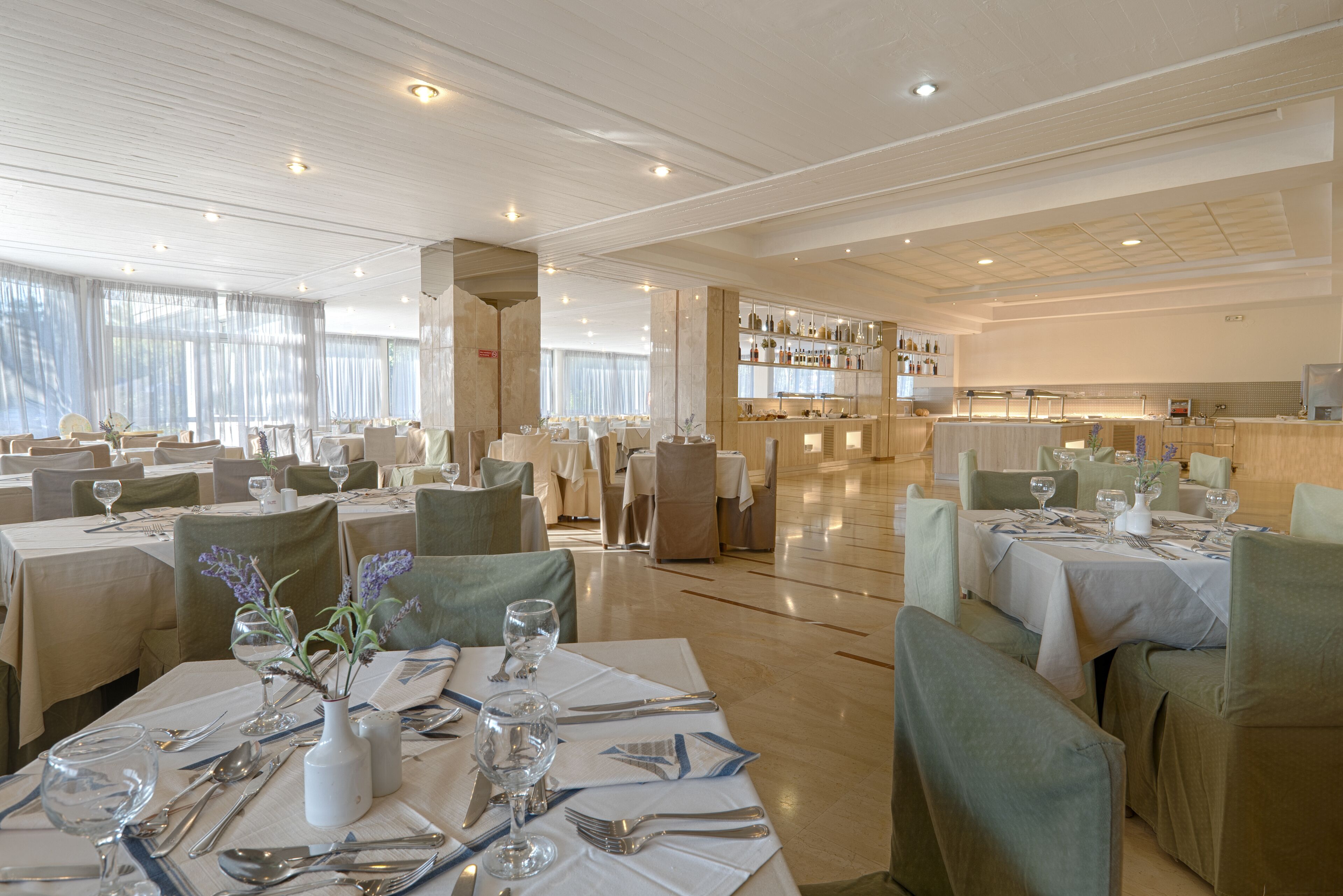 A spacious restaurant with multiple dining tables, each set with cutlery and glassware, and a bar area with shelves of bottles.