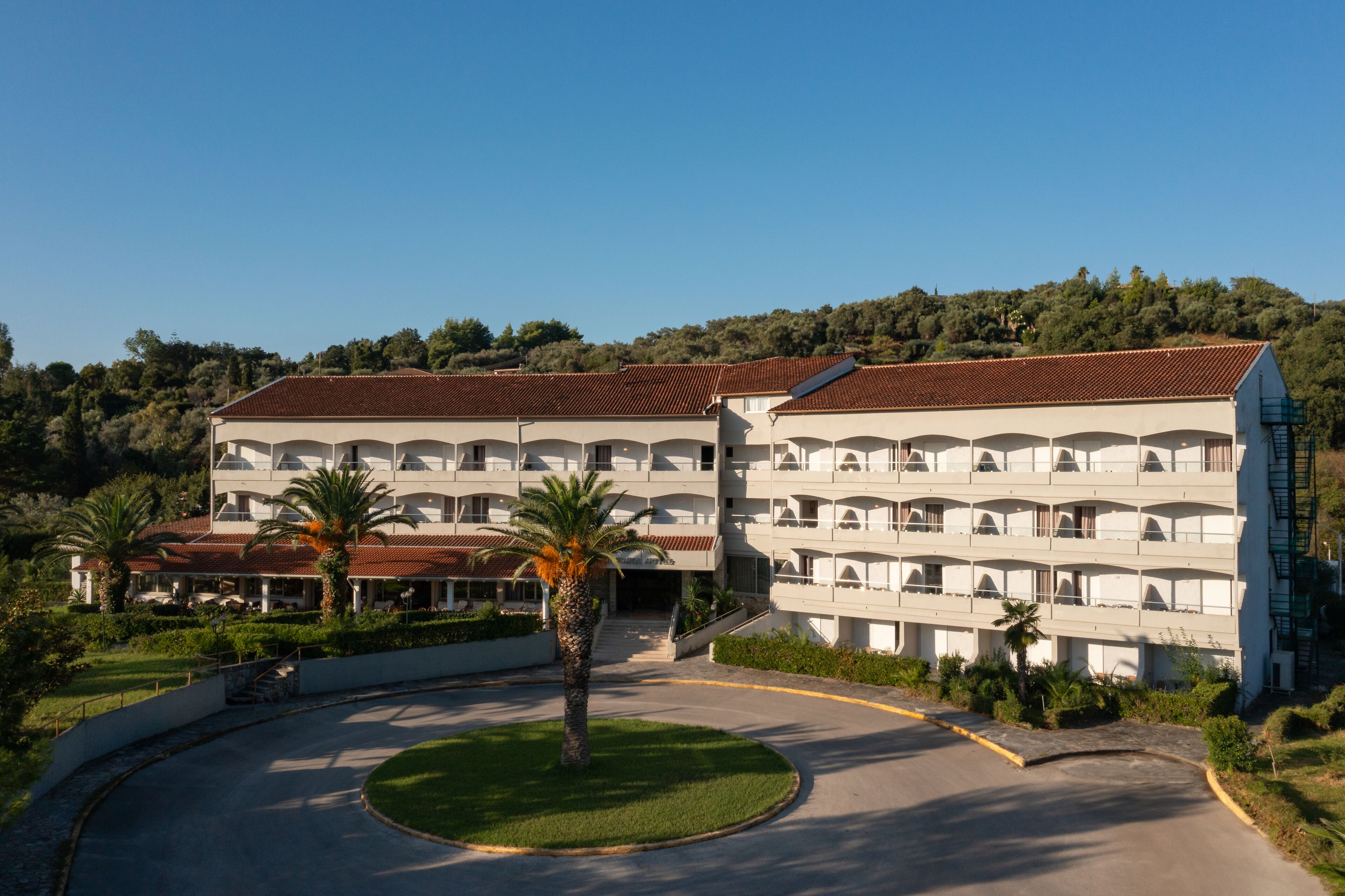 A large, multi-story hotel with a central courtyard, surrounded by lush greenery and a hilly backdrop.
