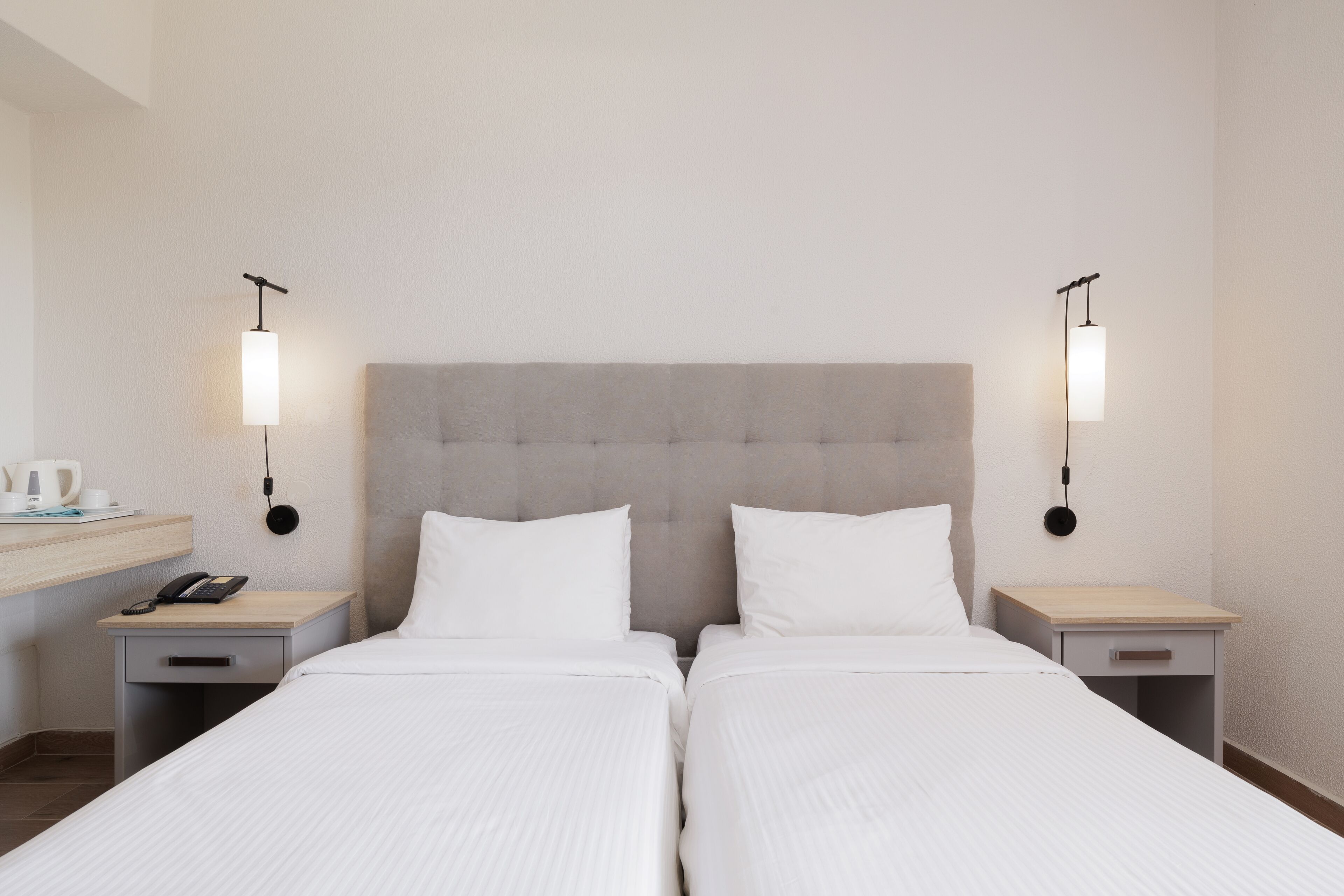 A hotel room with two single beds, a grey upholstered headboard, bedside tables with lamps, and a small desk with a telephone.