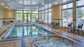 Una piscina interior con jacuzzi, rodeada de sillas y mesas, con ventanas amplias y un techo con iluminación empotrada.