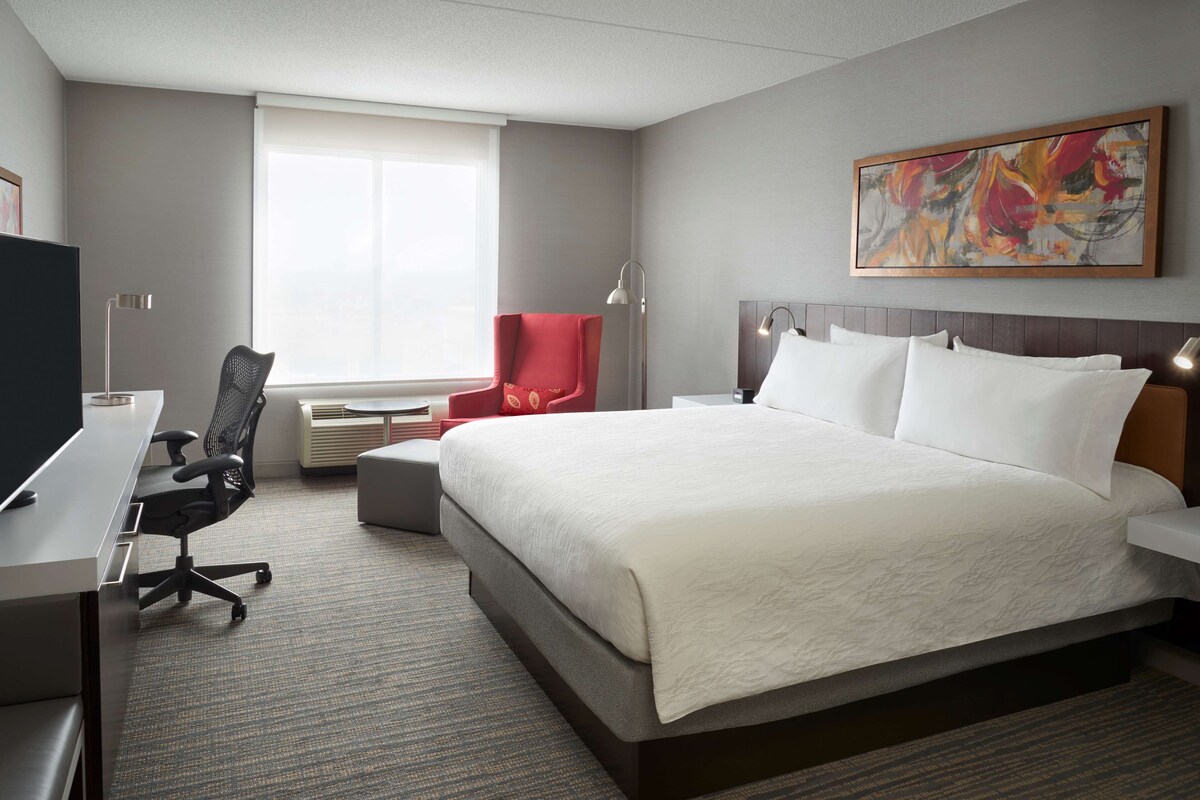 Habitación de hotel con una cama grande, un escritorio con silla, un sillón rojo y un cuadro en la pared.