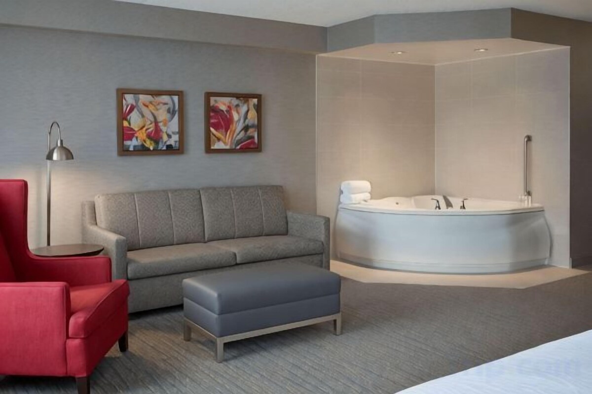 A modern hotel room with a grey sofa, a red armchair, a grey ottoman, a large bathtub, and two abstract paintings on the wall.