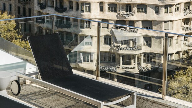 Terraza en la azotea con balaustrada de vidrio, una tumbona y vistas a un edificio histórico con balcones y esculturas.