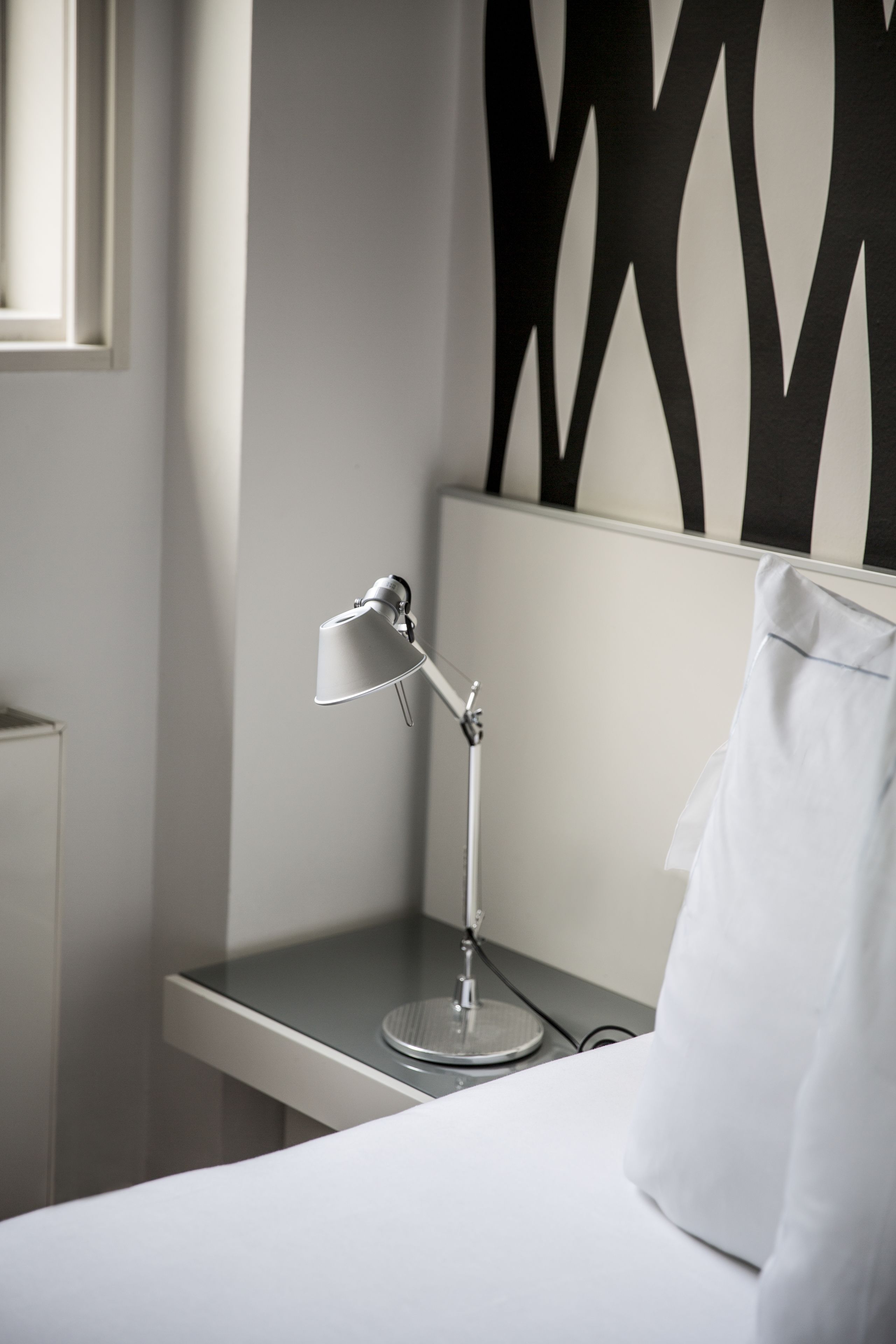 Una cama bien tendida con un almohadón blanco, una mesita de noche con una lámpara y una pared con un diseño geométrico.