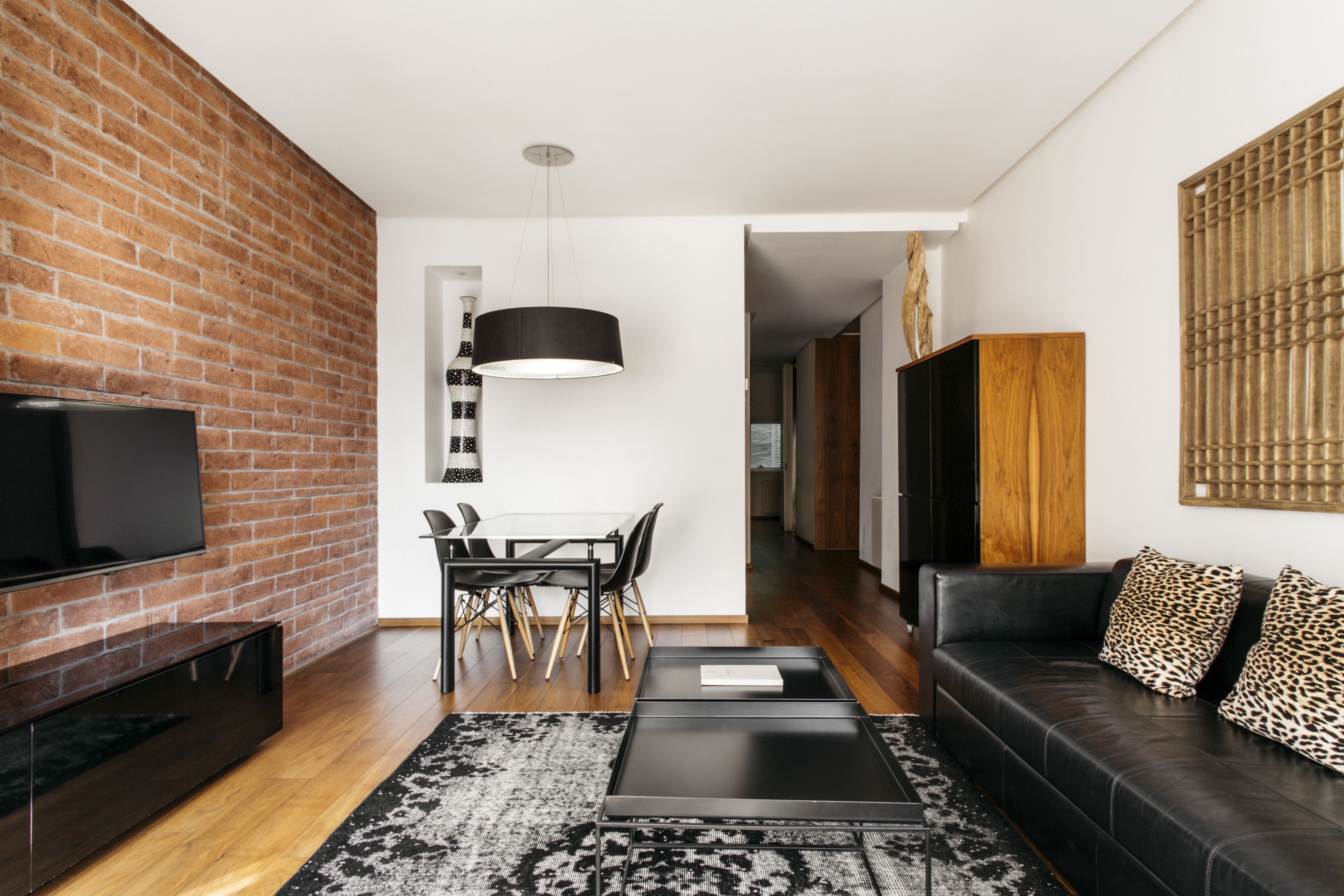 A modern living room with a brick wall, a flat-screen TV, a dining area with a table and chairs, a black leather sofa with leopard print cushions, and a black coffee table.