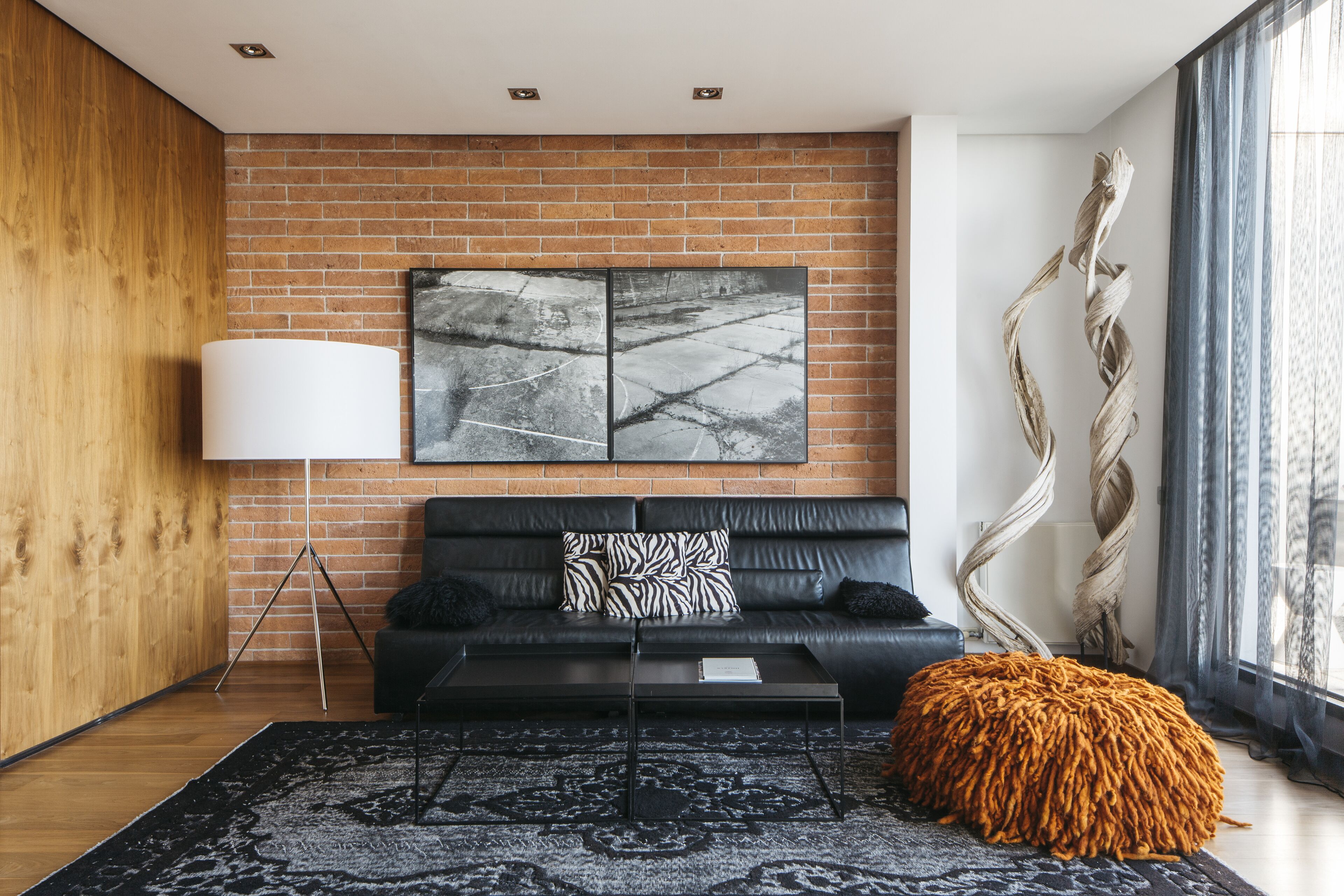 A modern living room with a brick wall, a black leather sofa, a white floor lamp, a patterned rug, and a large window with curtains.