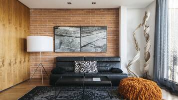 A modern living room with a brick wall, a black leather sofa, a white floor lamp, a patterned rug, and a large window with curtains.