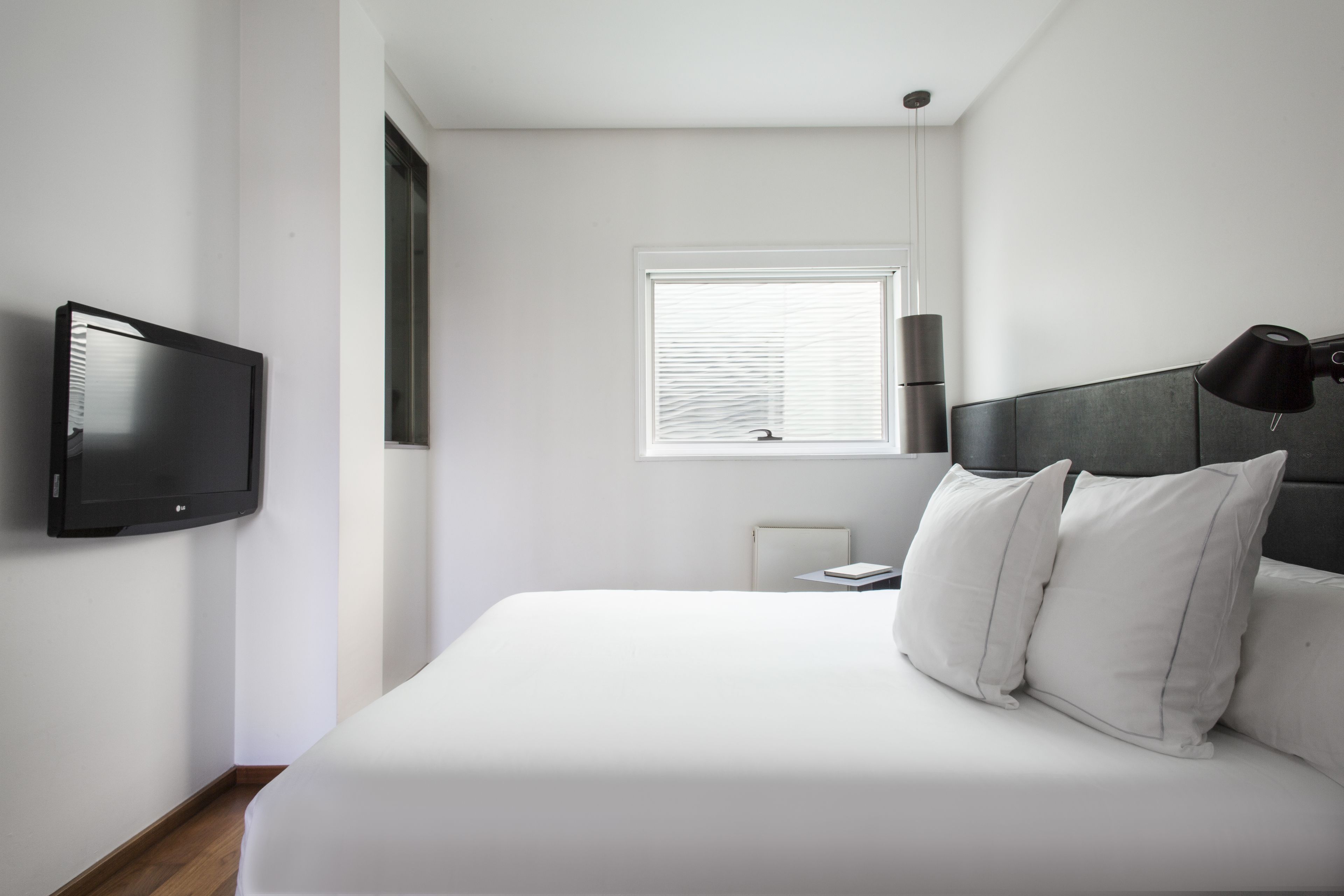 A modern bedroom with a bed, a TV mounted on the wall, a window with blinds, and a bedside lamp.