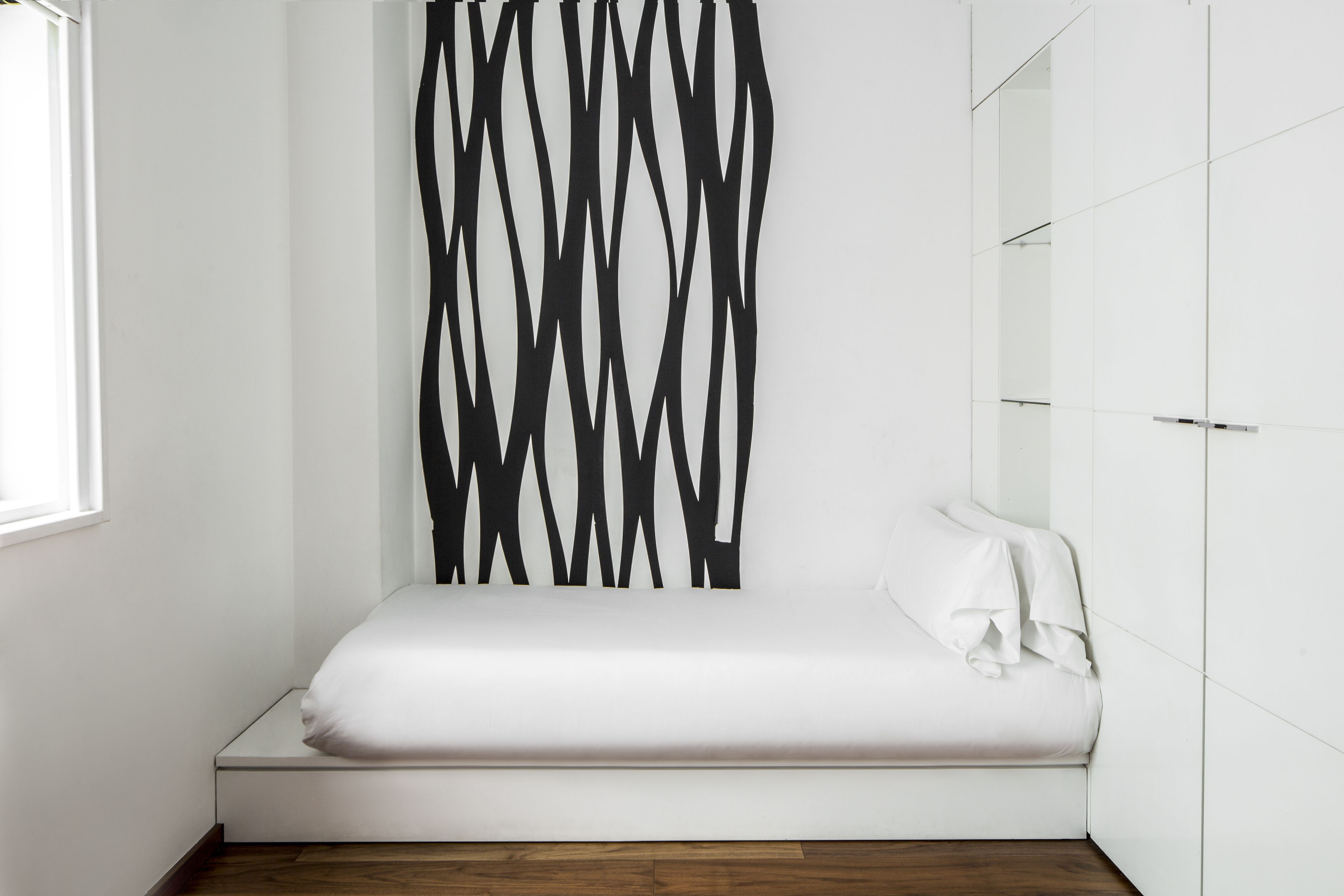 Una habitación pequeña y blanca con una cama individual, diseño moderno, un espejo grande en una pared y una ventana que deja entrar luz natural.