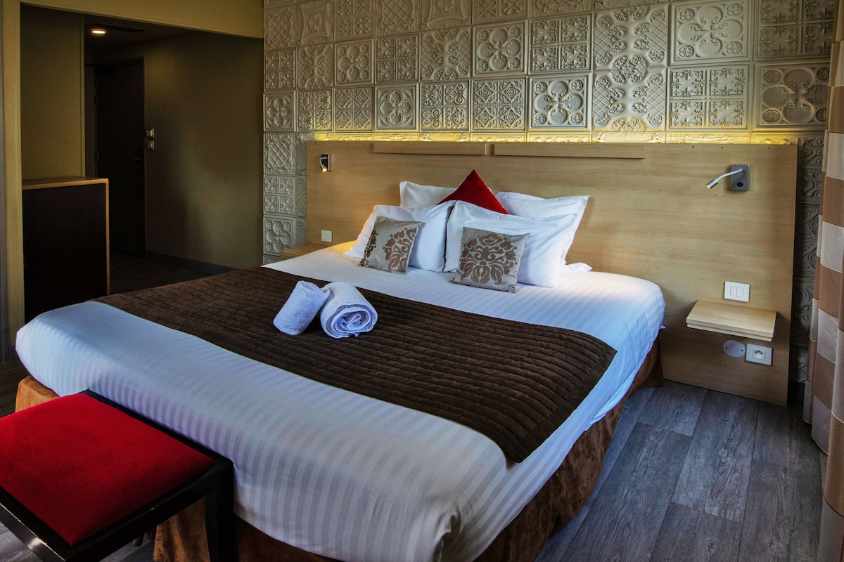 A neatly made bed with white and brown linens, a red pillow, and a brown throw blanket.