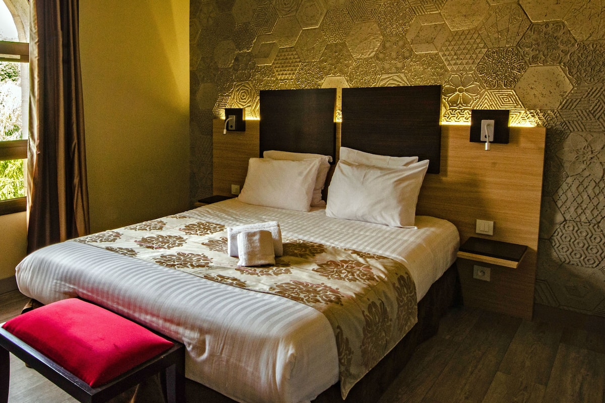A hotel room with a large bed, two bedside tables, a nightstand, a red bench, and a patterned wall.