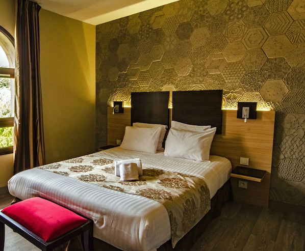 Habitación de hotel con una cama grande, dos mesitas de noche, un banco rojo y una pared estampada.