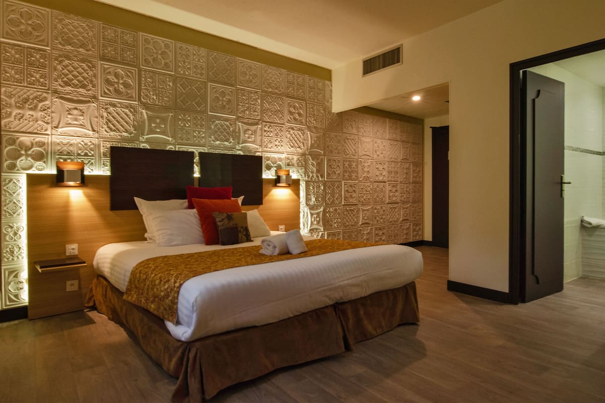 A hotel room with a large bed, a headboard with intricate patterns, a nightstand, and a wall-mounted lamp.