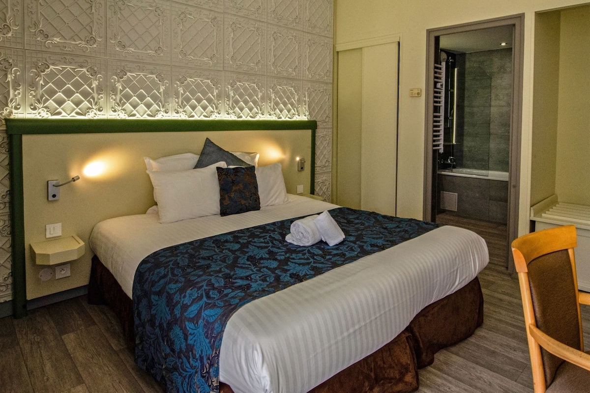 A hotel room with a bed, a nightstand, a chair, and a bathroom visible through an open door.
