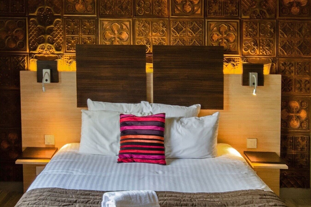 A neatly made bed with pillows, a blanket, and a folded towel. There are two bedside tables with lamps. The wall behind the bed has a decorative panel.